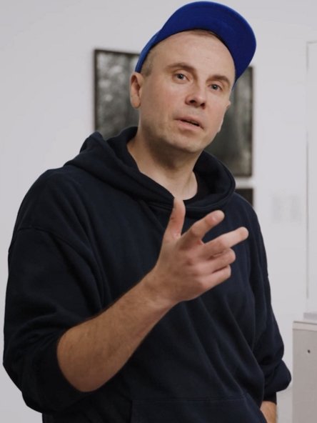 Filmstill: Eine Person mit dunkelblauer Kappe und schwarzem Pullover steht vor einer Ausstellungsvitrine mit Fotografien und gestikuliert mit einer Hand beim Sprechen.