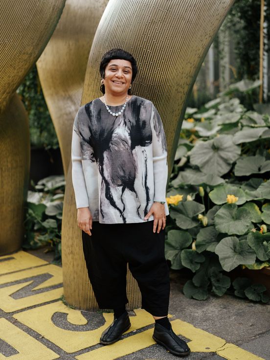 Foto: Porträt der Künstlerin Monira Al Qadiri vor einer silbernen Skulptur auf dem Vorplatz der Berlinischen Galerie. Sie blickt direkt in die Kamera und lacht.