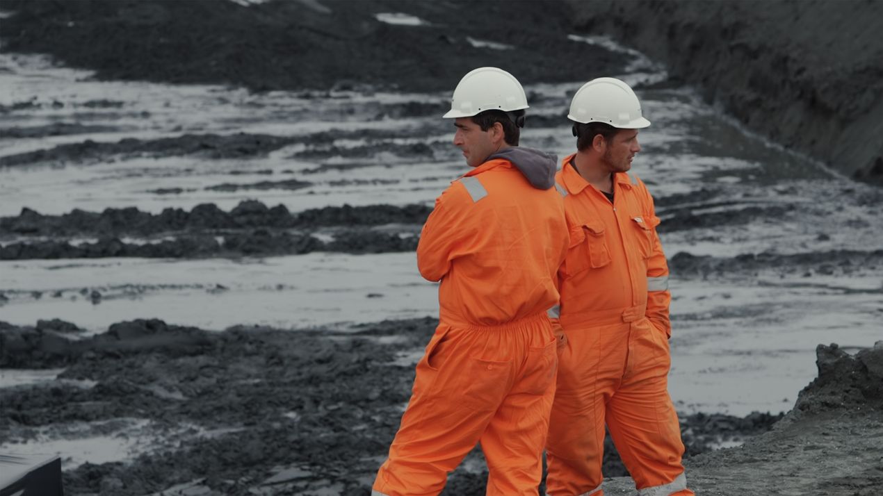 Film still: Two people in bright orange work clothes and white helmets stand in front of a building pit, their heads and bodies turned in different directions.