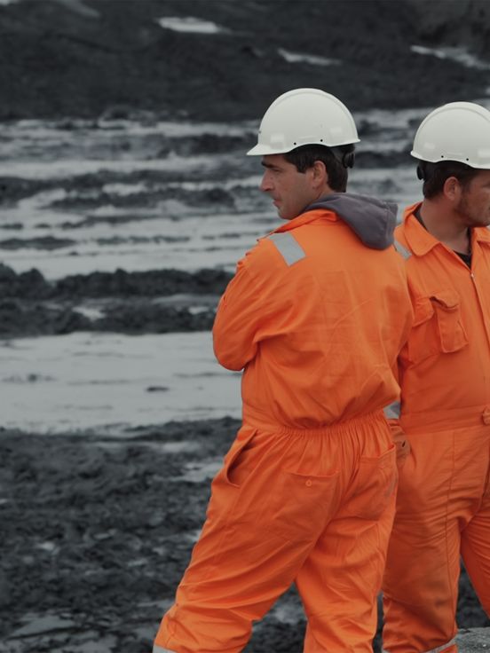 Film still: Two people in bright orange work clothes and white helmets stand in front of a building pit, their heads and bodies turned in different directions.