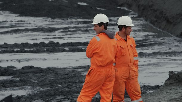 Film still: Two people in bright orange work clothes and white helmets stand in front of a building pit, their heads and bodies turned in different directions.