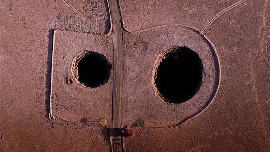 Luftaufnahme einer trockenen Landschaft mit zwei großen, kreisförmigen Löchern im Boden. Ein Weg führt in gerader Linie zwischen den Löchern hindurch und teilt die Fläche symmetrisch. Um die Löcher und den Weg herum sind Muster und Spuren im Boden sichtbar, die möglicherweise von Fahrzeugen oder anderen Aktivitäten stammen.