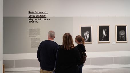 Foto: Drei Menschen im Ausstellungsraum. An der Wand Fotoporträts und die der Ausstellungstitel "Kann Spuren von Umbo enthalten".