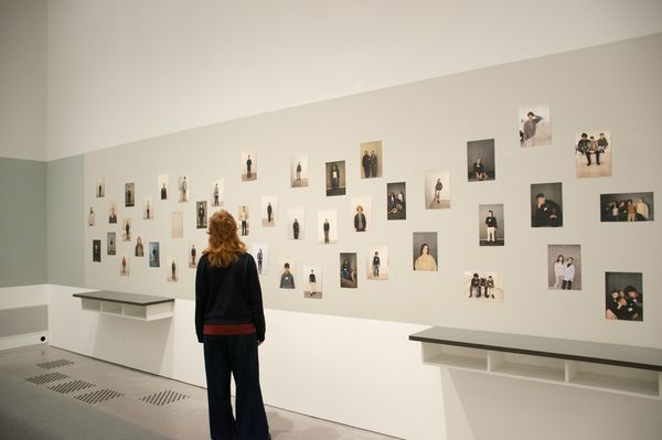 eine Person steht vor einer Wand, auf der in einer Peterburgerhängung Fotoportraits von Schüler*innen zu sehen sind