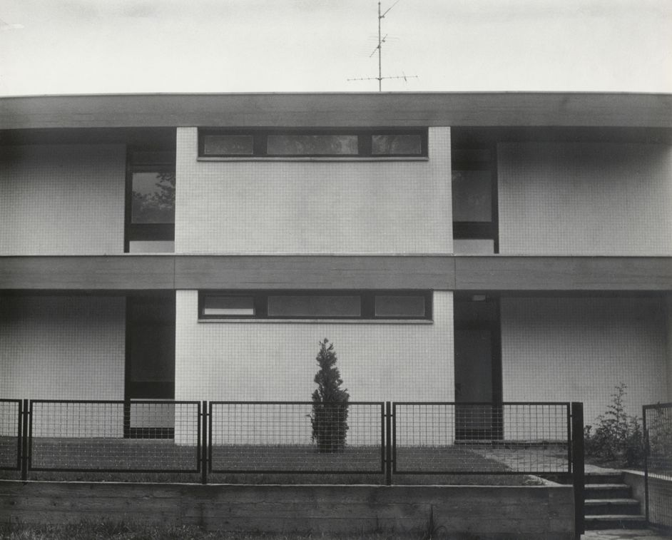Black and white photograph: Frontal view of a modern house façade with a flat roof and black metal garden fence.