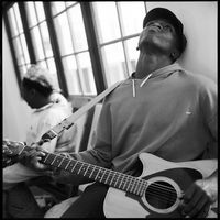 Square black and white photograph: In the foreground, a male figure wearing a hoodie can be seen playing the guitar and looking up towards the ceiling. Another person can be seen slightly blurred in the background. Both are sitting in front of a long window.