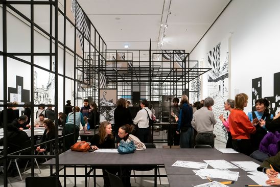 Photo: View into the exhibition space of "Kotti-Shop/SuperFuture" with many people, two long tables and a large lattice construction.