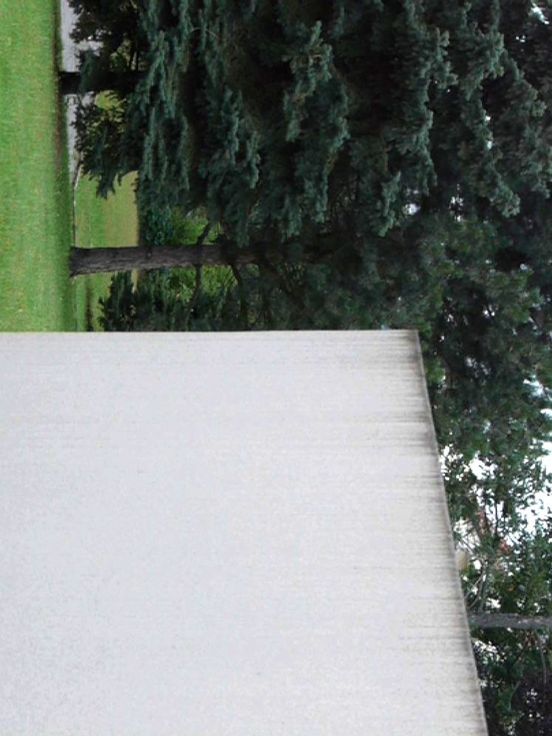 Film still: View of a paved path, a white wall surrounded by grass and trees behind it. The image is actually a portrait format, but has been rotated to the right.