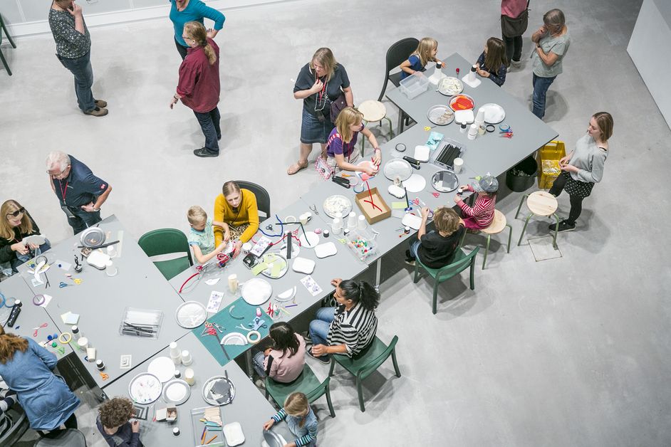 Photo: Children and adults stand and sit around a long table with many craft materials. Some are crafting together, others are talking.