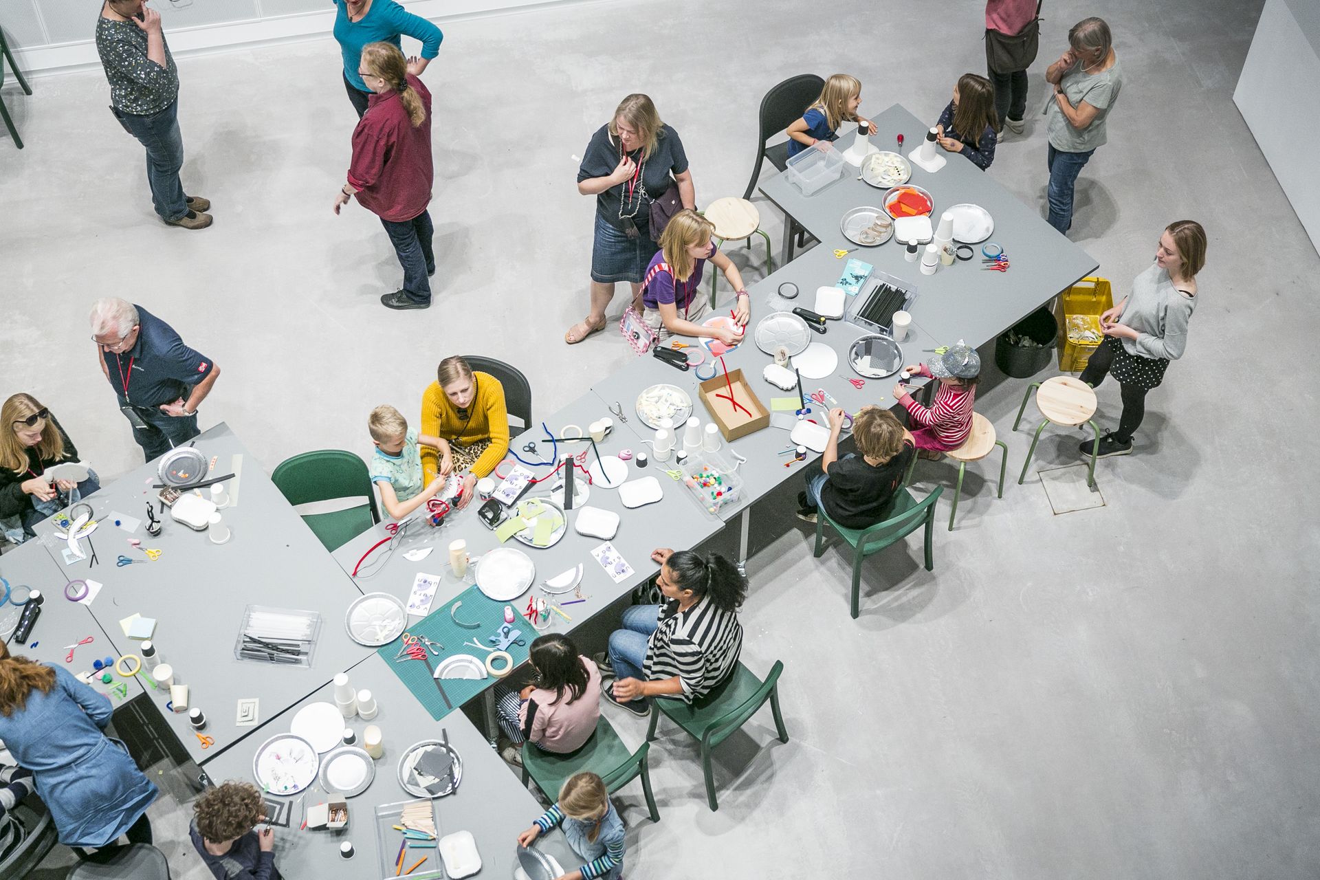 Familien*Tag im Raum 207 m² am 14.9.2019 Foto: Kinder und Erwachsene stehen und sitzen um einen langen Tisch mit vielen Bastelmaterialien. Manche basteln gemeinsam, andere unterhalten sich.
