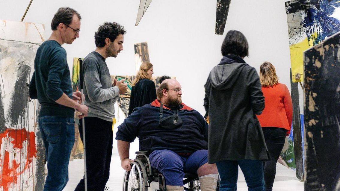 Foto: Menschen bewegen sich in einer Rauminstallation. Eine Person mit Rollstuhl, eine Person mit Blindenlangstock.
