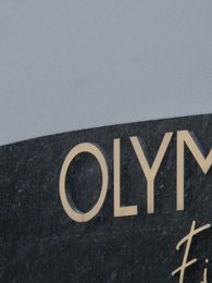 Filmstill: Das Still zeigt angeschnitten die Vorderseite des Olympia Einkaufszentrums in München und einen Teil des Schriftzugs an der Gebäudefassade in goldenen Lettern.