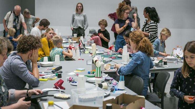 Foto: Viele Kinder und Erwachsene stehen und sitzen um einen L-förmigen Tisch mit vielen Bastelmaterialien. Manche basteln gemeinsam, andere unterhalten sich.