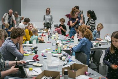 Foto: Viele Kinder und Erwachsene stehen und sitzen um einen L-förmigen Tisch mit vielen Bastelmaterialien. Manche basteln gemeinsam, andere unterhalten sich.