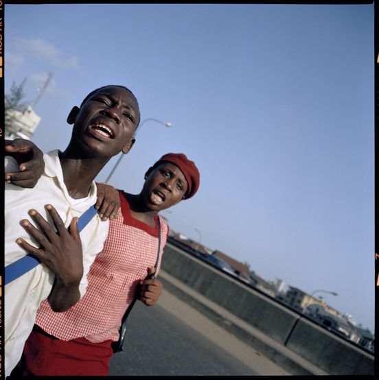 Square color photograph: The photograph shows two people in the foreground, a younger, male person and to the right an older, female person. A street, lanterns and houses can be seen in the background. Next to the two people, the rich, light blue sky takes up a large part of the picture.