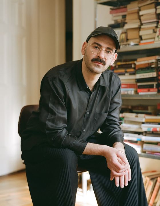 Foto: Enrico Ippolito sitzt in einem Innenraum auf einem Stuhl und blickt direkt in die Kamera. Der Hintergrund zeigt links eine weiße Tür und rechts ein volles Bücherregal.