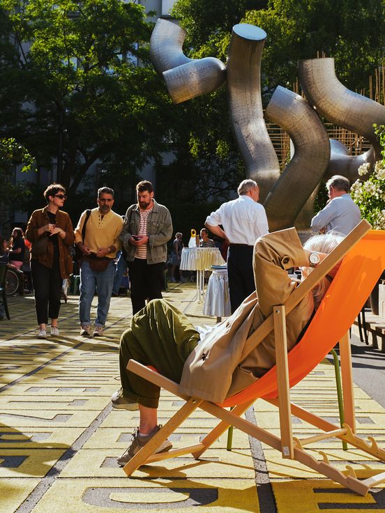 Foto: Blick auf den belebten Vorplatz der Berlinischen Galerie. Links vorne sitzt eine Person mit über dem Kopf verschränkten Armen gemütlich in einem orangenen Liegestuhl, im Hintergrund laufen Menschen über den Platz.