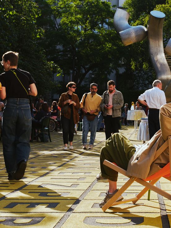Photo: View of the busy forecourt of the Berlinische Galerie. In the foreground on the left, a person sits comfortably in an orange deckchair with his arms crossed over his head; in the background, people walk across the square.