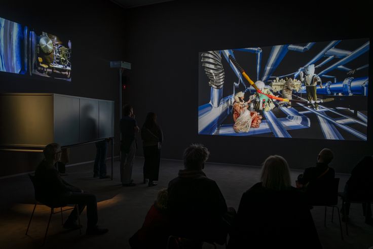 Ausstellungsansicht „Psychonauten. John Bock und Heiner Franzen“, Berlinische Galerie Foto: Ein paar Menschen sitzen und stehen in einem Videoraum und schauen auf eine große Leinwand.