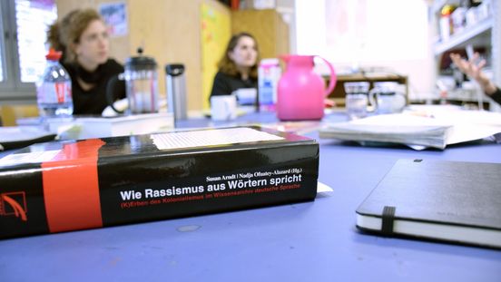 Foto: An einem Tisch sitzen zwei Menschen an einem Tisch mit Arbeitsmaterialien. Im Vordergrund ein Buch mit dem Titel "Wie Rassismus aus Wörtern spricht"