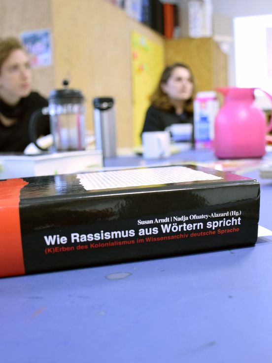 Foto: An einem Tisch sitzen zwei Menschen an einem Tisch mit Arbeitsmaterialien. Im Vordergrund ein Buch mit dem Titel "Wie Rassismus aus Wörtern spricht"