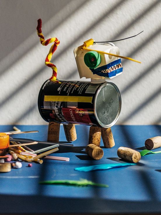 Foto: Eine hunde-ähnliche kleine Skulptur aus verschiedenen, bunter Materialien steht auf einem blauen Untergrund. Um sie herum verteilt liegen verschiedene Gegenstände, wie Scheren, Korken und Strohalme.