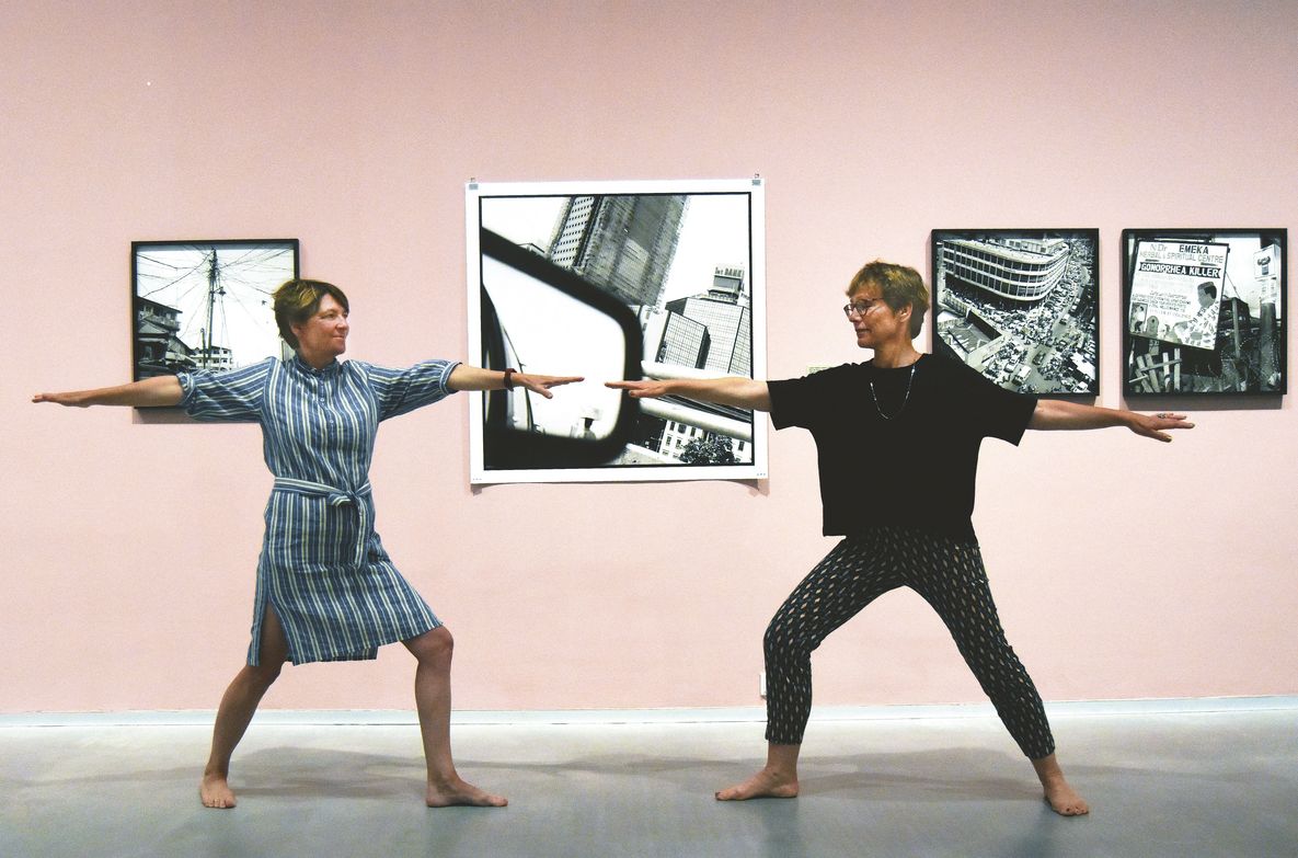 Foto: Zwei Personen stehen sich in Yoga-Pose gegenüber und blicken einander an. Im Hintergrund sind schwarz-weiß-Fotografien von Akinbode Akinbiyi vor einer rosafarbenen Wand zu sehen.