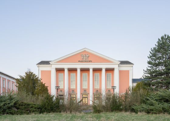 Frontalansicht eines rosafarbenen Kulturhauses mit weißem Säulenportikus, umgeben von Bäumen und Sträuchern unter klarem Himmel