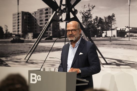 Photo: Thomas Köhler stands at a lectern in front of a photo wallpaper in black and white. The wallpaper shows a sculpture by Hans Uhlmann at Hansaplatz in Berlin.