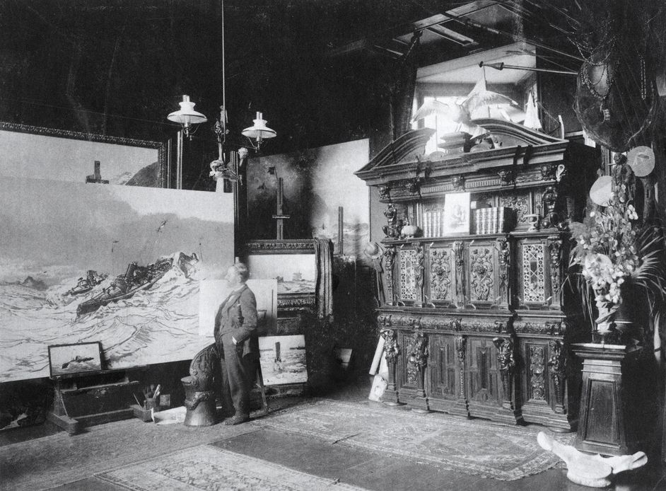 Schwarz-Weiß-Fotografie: Mann steht in einem Atelier vor Gemälden auf Staffeleien