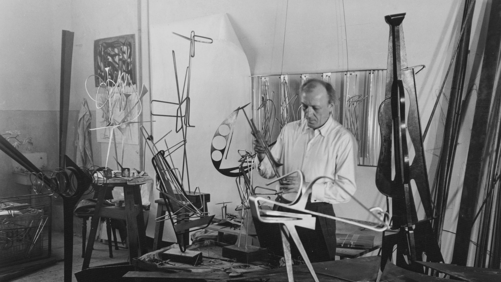 Schwarz-Weiss-Fotografie von Hans Uhlmann in seinem Atelier. Um den Künstler herum sind viele Metall-Skulpturen im Entstehungsprozess zu sehen.