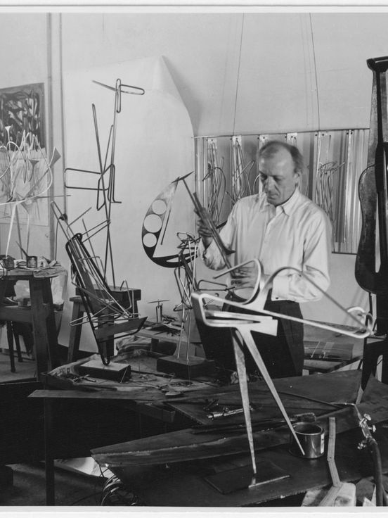 Schwarz-Weiss-Fotografie von Hans Uhlmann in seinem Atelier. Um den Künstler herum sind viele Metall-Skulpturen im Entstehungsprozess zu sehen.