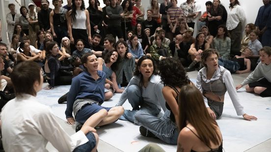 Foto: Einige Performer*innen sitzen in unterschiedlichen Positionen auf dem Boden, umringt von Publikum.