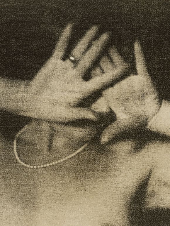 Grainy black-and-white photograph: Portrait of a nude person wearing a pearl necklace, holding both hands in front of their face, making it unrecognizable. The hands are adorned with jewelry, including a ring and a bracelet.