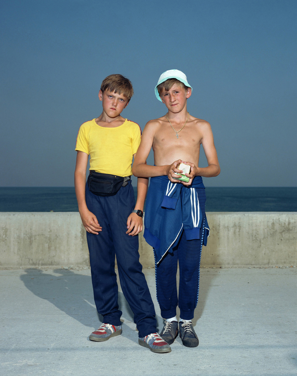 Farb-Fotografie: Zwei Jungen stehen vor einer Beton-Ballustrade, im Hintergrund ist das dunkelblaue Meer zu sehen. Darüber erhebt sich ein wolkenloser, blauer Himmel. Beide Jungen tragen Trainingshosen und blicken direkt in die Kamera.