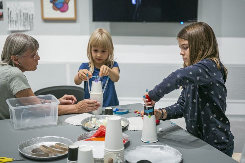 Foto: Zwei Kinder und eine Erwachsene sitzen um einen Tisch mit Bastelmaterialien. Die Kinder basteln Gebilde aus Pappbechern.