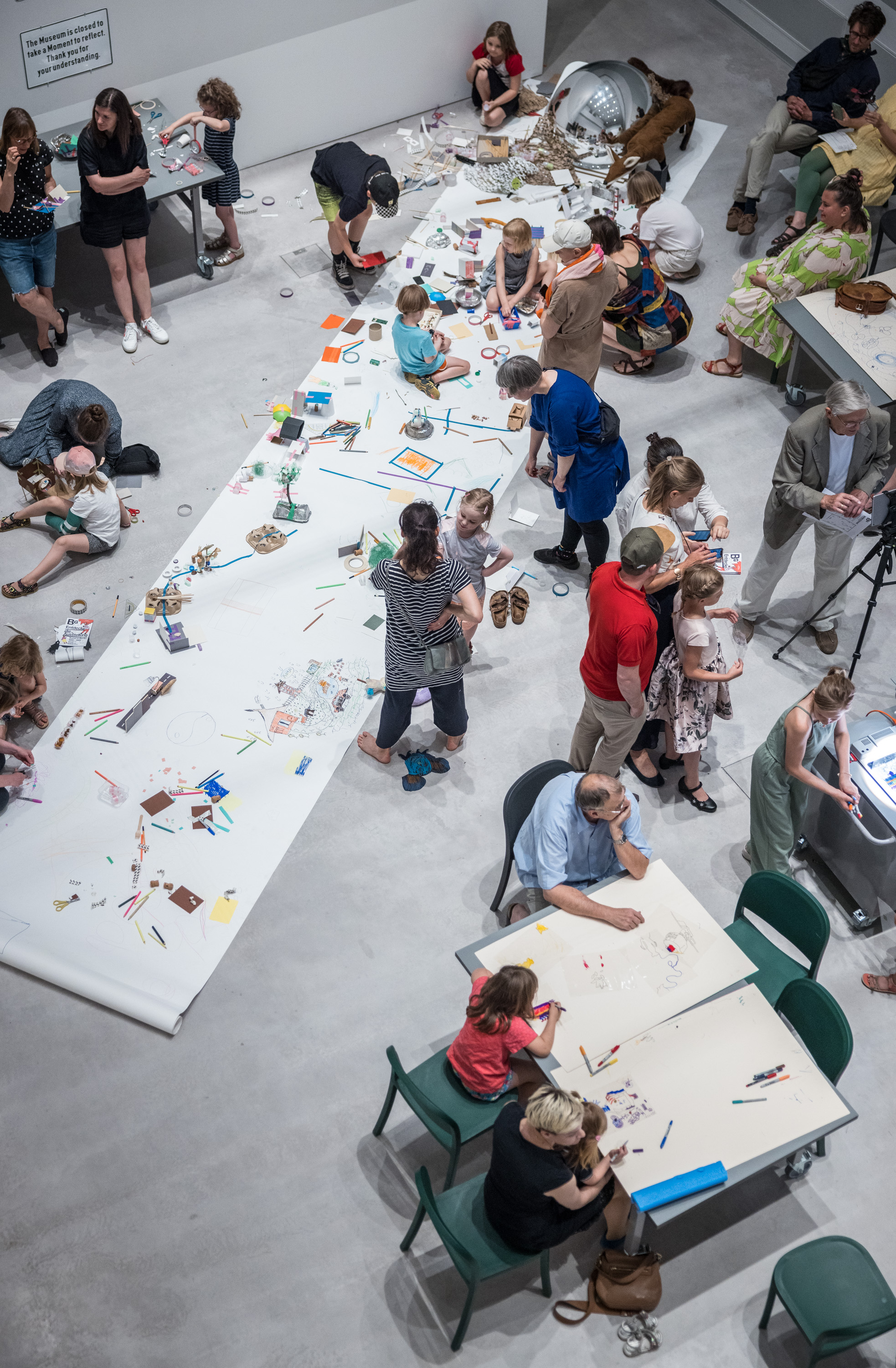 Foto in einen Raum mit vielen Kindern und Erwachsenen, die an Tischen und einer großen Papierbahn stehen und sitzen