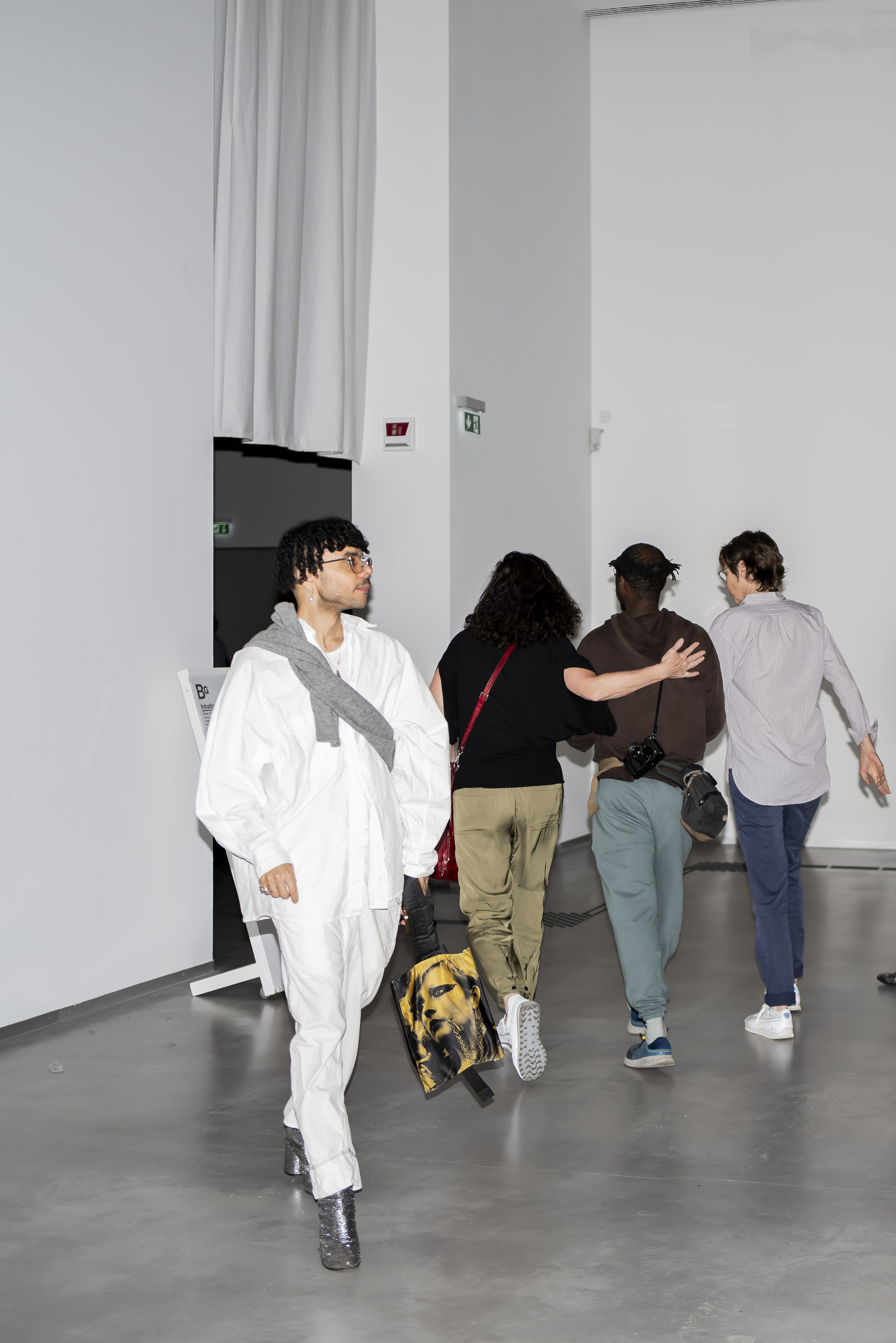 Photo with flash: Front view of a person in white clothing and glittering silver shoes walking out of an exhibition room, while three people walk into the room in the background.